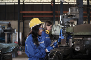 An Industrial workers team in protective and safety uniforms and hardhats, male manager, and female colleagues work with metalwork machines in manufacturing factory. Professional production engineer.