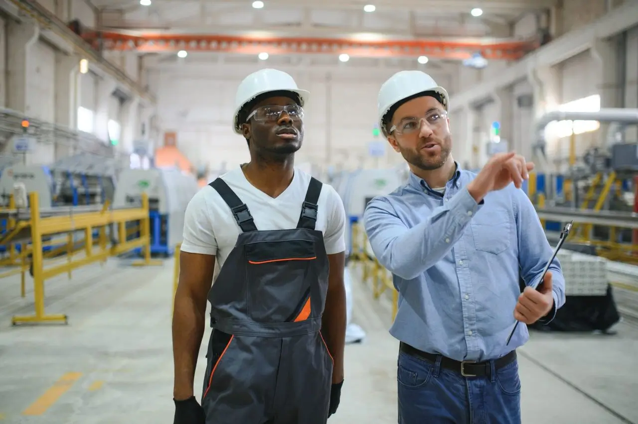 Two workers wearing hard hats engaged in discussion while standing inside a warehouse